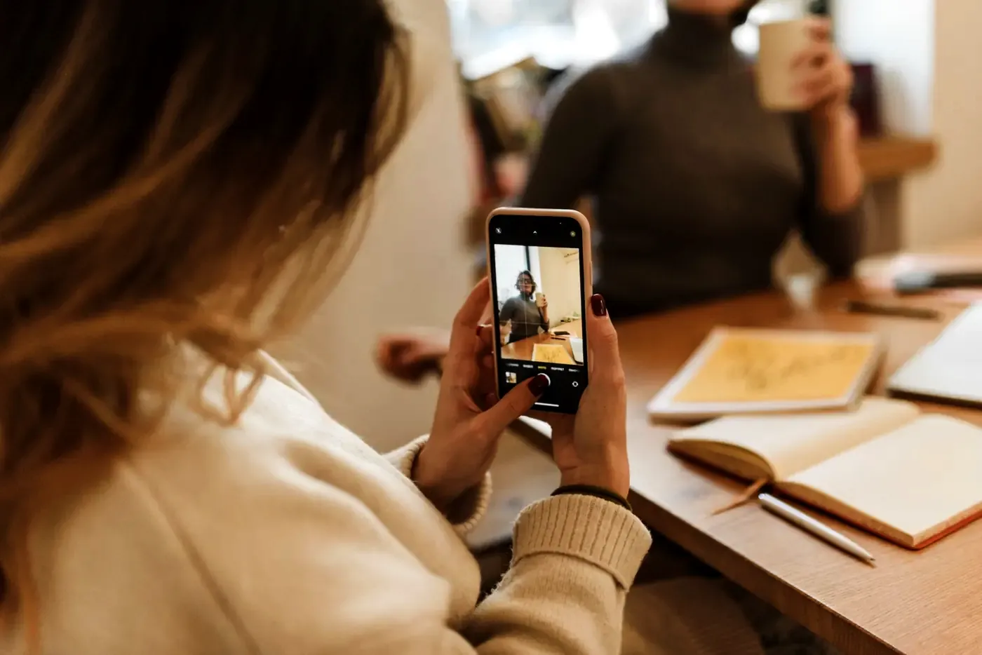Një grua fotografon me telefon një bashkëbiseduese në tavolinë pune